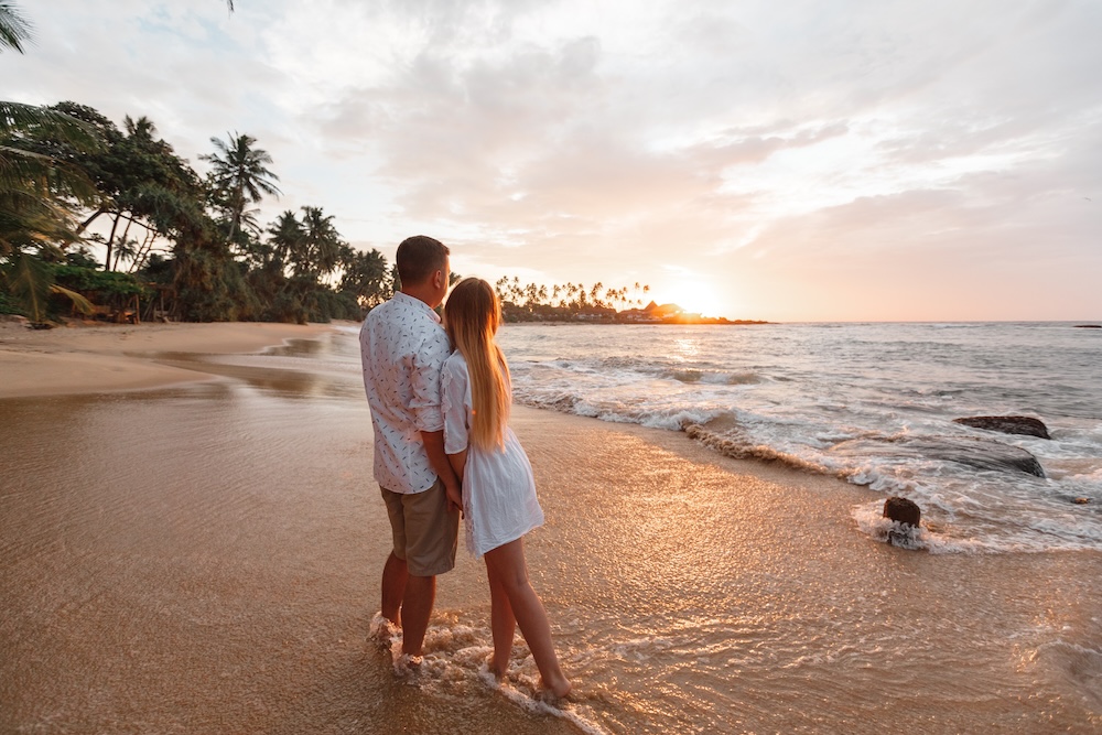 Young happy couple on seashore, looking beautiful golden sunset during vacation on tropical islands, palm trees on background, waves touch their feets.